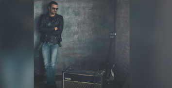 man leaning on wall behind VOX amplifier and beside electric guitar